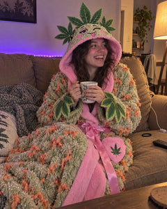 Woman curled up on a couch in the Wake & Cozy robe, hood up, holding a mug